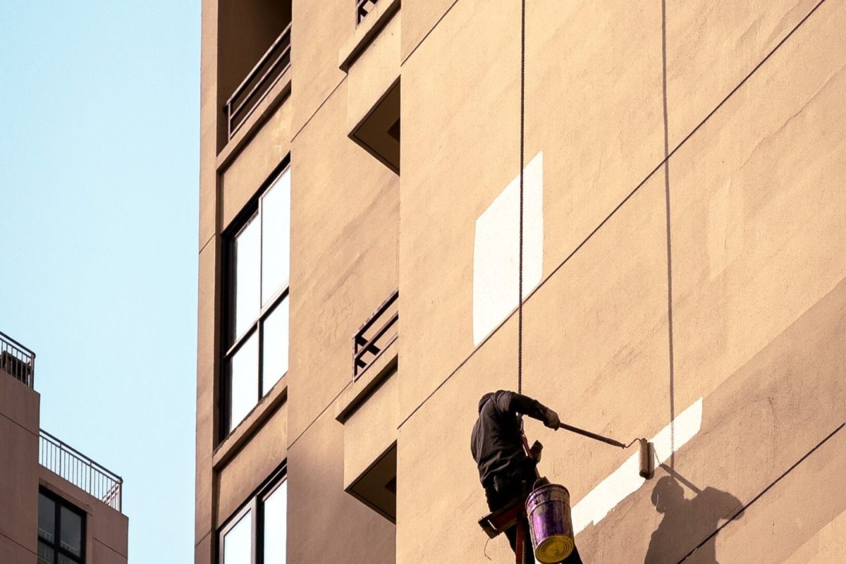 Man painting tall building exterior while suspended outdoors, showcasing urban maintenance work.