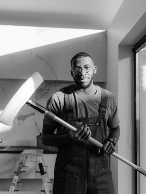 Black and white image of a male worker in overalls holding a paint roller indoors.